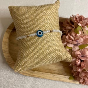 Könnte beinhalten: Ein weißes Perlenarmband mit einem blauen und weißen Evil-Eye-Anhänger und goldenen Akzenten. Das Armband wird auf einem Jutekissen und einem Holztablett mit rosa Blumen im Hintergrund präsentiert.