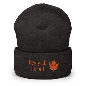 May include: Dark gray beanie with the embroidered text "hey y'all its fall" and an orange maple leaf. The beanie is a fall-themed accessory, perfect for cooler weather. The hat is made of a soft material.
