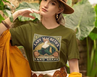 Op de afbeelding: Een olijfgroen t-shirt met een grafische print van een vrouw in een donkergroene jurk met de tekst Lady Macbeth's Perfumes of Arabia & Hand Soaps. Het ontwerp is omlijst door een rechthoekige rand met decoratieve florale elementen.