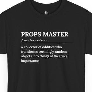 May include: A black t-shirt featuring white text that reads PROPS MASTER, followed by the phonetic pronunciation /præps ˈmæstər/ noun, and the definition: A collector of oddities who transforms seemingly random objects into things of theatrical importance.