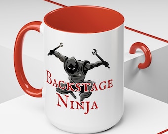 May include: White ceramic mug with a red interior and handle. The mug features a graphic of a ninja holding wrenches, with the words "Backstage Ninja" in red. The mug is designed for beverages.