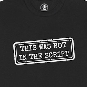 Puede incluir: Camiseta negra con un gráfico rectangular blanco que dice "THIS WAS NOT IN THE SCRIPT". La camiseta tiene cuello redondo y un pequeño logotipo cerca del cuello. El texto está en negrita, fuente sans-serif.