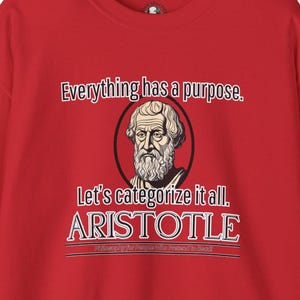 Puede incluir: Camiseta roja con un gráfico blanco de Aristóteles, un filósofo griego, con el texto "Todo tiene un propósito. Categoricemos todo. ARISTÓTELES Filosofía para la gente que finge leer".
