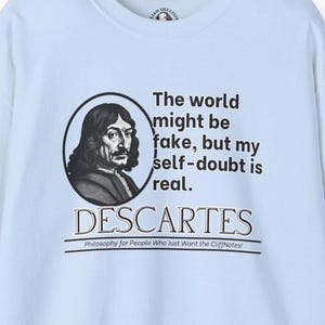 Puede incluir: Camiseta azul claro con una ilustración en blanco y negro de René Descartes, un famoso filósofo. El texto dice "El mundo podría ser falso, pero mi duda de mí mismo es real. DESCARTES Filosofía para la gente que solo quiere las CliffNotes!"