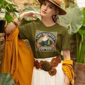 May include: An olive green t-shirt featuring a graphic print of a woman in a green dress with the text Lady Macbeth's Perfumes of Karma & Hand Soaps, paired with a white skirt, a wide brown circular metal belt, and a tan felt fedora hat.