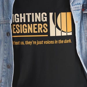 Puede incluir: Camiseta negra con las palabras "LIGHTING DESIGNERS" en letras blancas y doradas. La camiseta también presenta el texto "without us, they're just voices in the dark." También hay un gráfico de un sol o una luminaria.