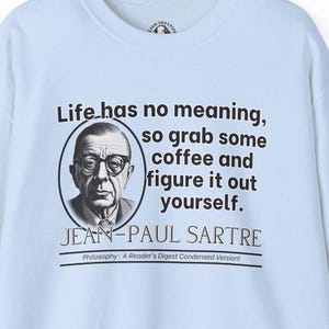 Puede incluir: Sudadera azul claro con una ilustración en blanco y negro de Jean-Paul Sartre. El texto dice "La vida no tiene sentido, así que toma un café y descúbrelo por ti mismo. Jean-Paul Sartre. Filosofía: ¡Una versión condensada de Reader's Digest!"