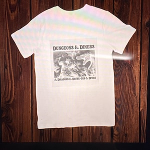 May include: White t-shirt with a graphic that reads "DUNGEONS & DINERS" above a black and white illustration of fantasy creatures. Below the illustration, the text reads "& DRAGONS & DRIVE-INS & DIVES". The shirt has short sleeves.