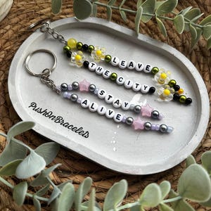 Könnte beinhalten: Schlüsselanhänger mit Perlendesign und den Sätzen "LEAVE THE CITY" und "STAY ALIVE" in weißen Buchstabenperlen. Die Schlüsselanhänger haben silberfarbene Hardware, transparente Daisy- und sternförmige Perlen und werden auf einem grauen Tablett präsentiert.
