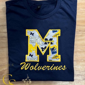 May include: A navy blue t-shirt with a large yellow embroidered "M" featuring a collage of Michigan Wolverines logos and the text "Wolverines" embroidered below.
