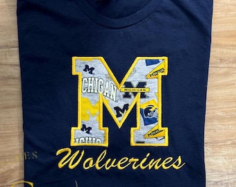 May include: A navy blue t-shirt with a large yellow embroidered "M" featuring a collage of Michigan Wolverines logos and the text "Wolverines" embroidered below.