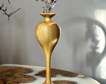 May include: A tall, wooden vase with a slender, teardrop-shaped body and a wide, flared opening. The vase is made of light-colored wood with a natural grain pattern. It is sitting on a table with a white and brown epoxy resin surface.