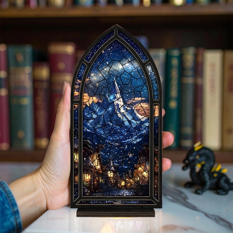 Może przedstawiać: Drewniana lampka nocna z łukowatym wierzchołkiem, prezentująca efekt witrażu. Projekt przedstawia gwiaździste niebo nad g&oacute;rą i sceną wioski. Rama i podstawa są z ciemnego drewna, z ciepłym światłem emanującym z wnętrza.