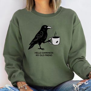 Puede incluir: Sudadera verde oliva con un cuervo negro sosteniendo una taza de café. El texto "Hello Darkness My Old Friend" está impreso debajo. Es una prenda informal de manga larga, adecuada para el uso diario.
