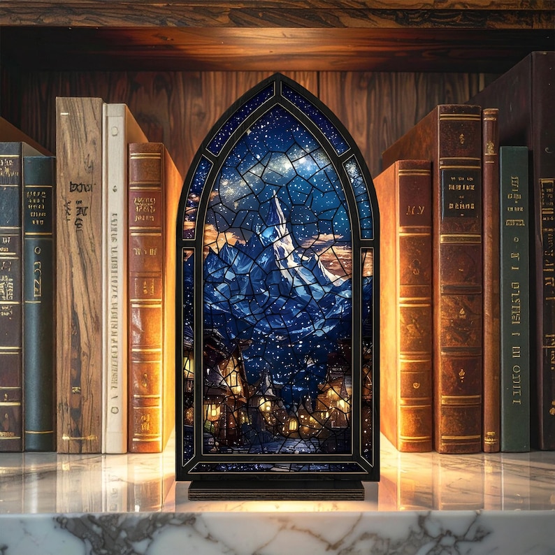 Może przedstawiać: Dekoracyjny witraż w kształcie gotyckiego łuku, przedstawiający nocną scenę z g&oacute;rą i wioską. Panel jest oprawiony w czarną ramę i stoi na marmurowej powierzchni, z antycznymi książkami w tle.