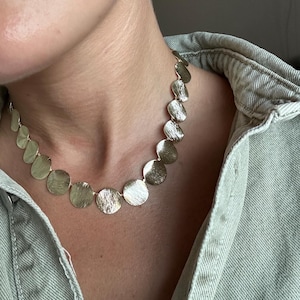 Könnte beinhalten: Eine goldfarbene Halskette mit strukturierten, kreisförmigen Anhängern. Die Halskette wird über einem hellgrünen Jeanshemd getragen. Die Anhänger sind durch eine zarte Kette verbunden, was einen modernen und eleganten Look erzeugt. Ein modisches Accessoire.