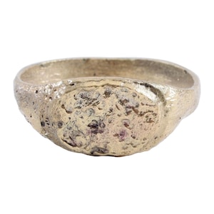 May include: A textured, silver-colored ring with an oval face. The ring has a rough, uneven surface, giving it a rustic appearance. The band is thick and sturdy, and the face of the ring has a slightly raised, textured design.