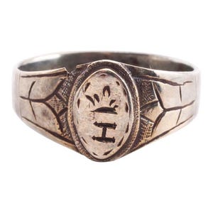 May include: A silver ring with an oval-shaped design featuring a crown and the letter "H". The ring has a textured band with intricate details.