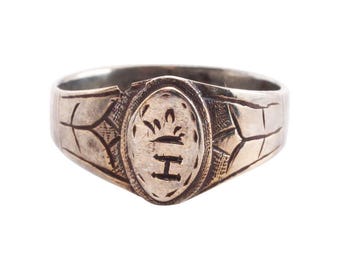 May include: A silver ring with an oval-shaped design featuring a crown and the letter "H". The ring has a textured band with intricate details.