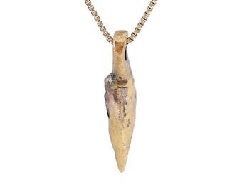 May include: A gold-colored pendant necklace with a unique, textured arrowhead-shaped pendant. The pendant hangs from a delicate chain, creating a simple yet striking accessory. The necklace is set against a white background.