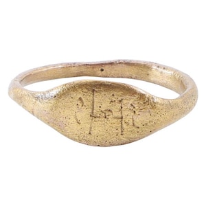 May include: A gold-toned metal signet ring with a textured, matte finish. The oval face of the ring features an engraved, linear symbol consisting of vertical and horizontal lines. The band has an irregular, organic shape with a slightly uneven surface.