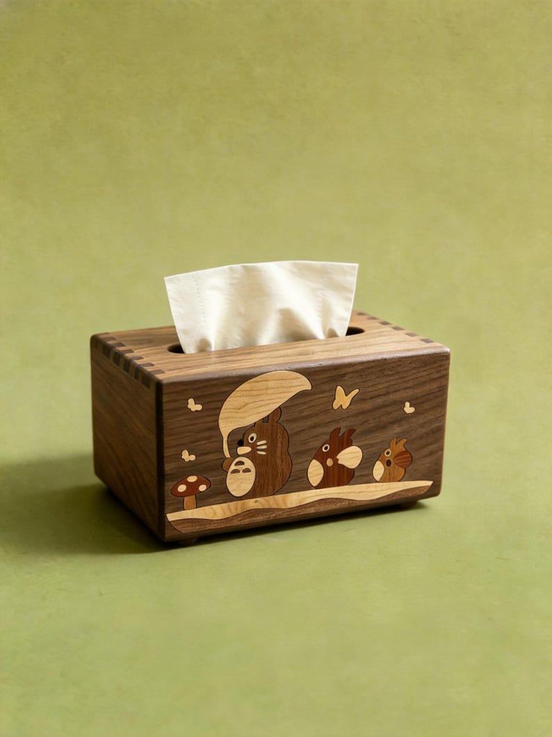 May include: A rectangular wooden tissue box holder featuring an inlaid wood design of three forest creatures, one holding an umbrella, alongside a small mushroom and butterflies. The box has visible finger joints at the corners and holds white tissues.