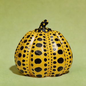May include: A small, rounded pumpkin sculpture featuring a bright yellow base colour with a pattern of solid black polka dots of varying sizes. The pumpkin has a short, curved stem at the top, also decorated with black dots on a yellow background.