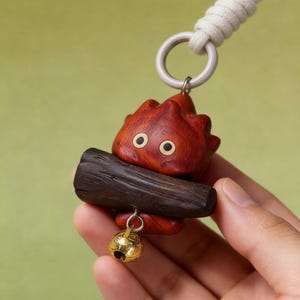 May include: A wooden pendant featuring a red, flame-shaped character with black eyes, holding a dark brown log. A small gold-colored bell hangs below. The pendant is attached to a white cord with a silver-colored ring.