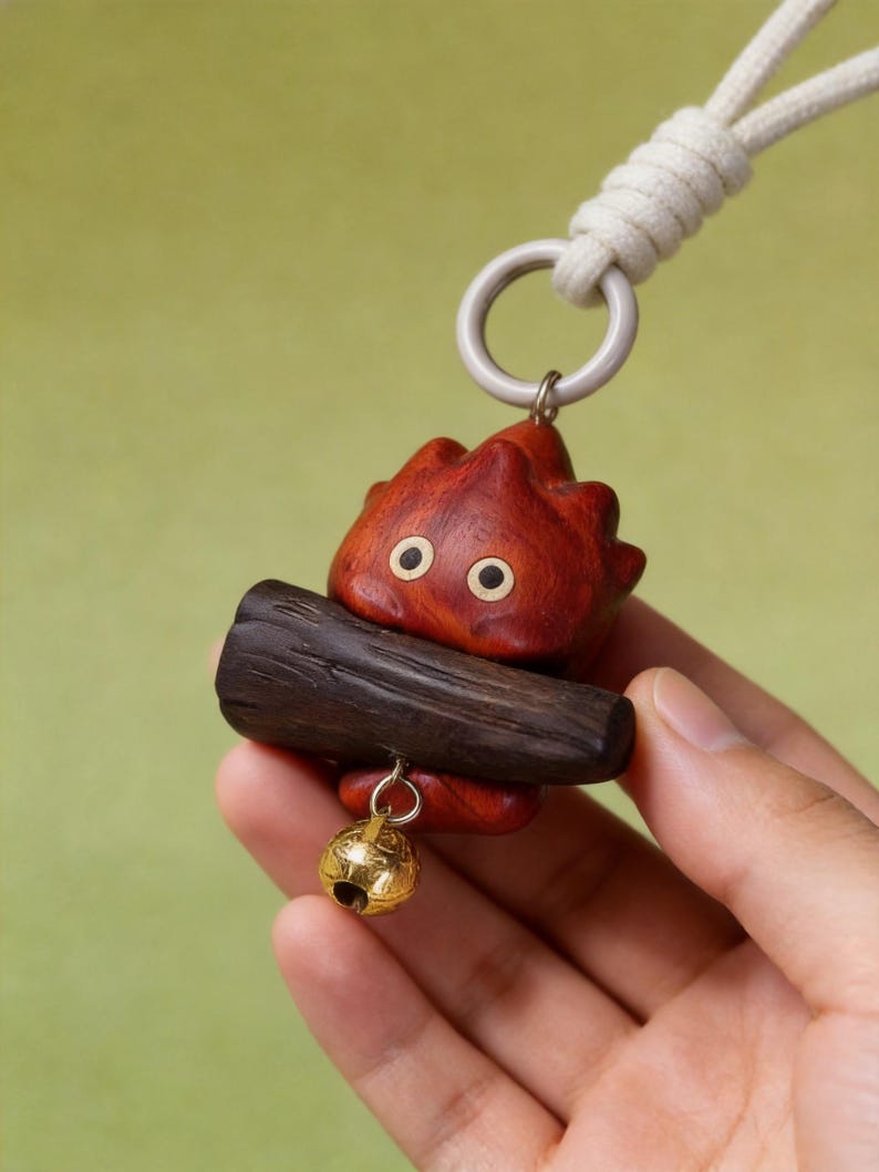 May include: A wooden pendant featuring a red, flame-shaped character with black eyes, holding a dark brown log. A small gold-colored bell hangs below. The pendant is attached to a white cord with a silver-colored ring.