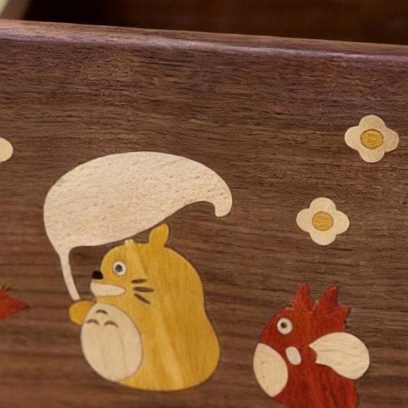May include: Wooden box with inlaid cartoon characters. A yellow creature with a white umbrella is featured, along with a red bird-like figure and small flower accents. The box has a dark brown wood grain finish.