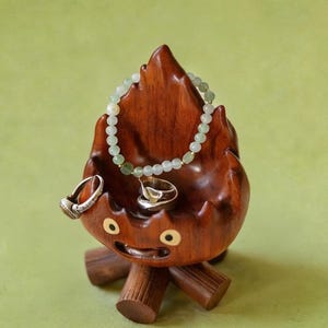 May include: A whimsical wooden jewelry holder shaped like a flame with a smiling face. It holds a beaded bracelet and two rings. The holder is brown with a light green background.