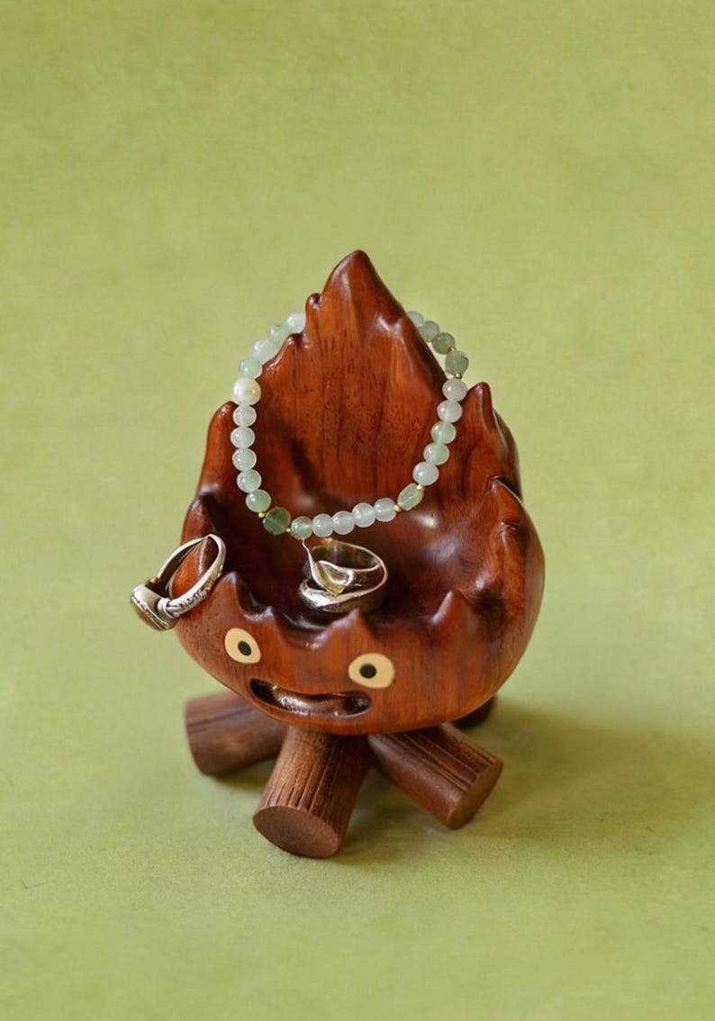 May include: A whimsical wooden jewelry holder shaped like a flame with a smiling face. It holds a beaded bracelet and two rings. The holder is brown with a light green background.