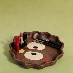 May include: A decorative wooden makeup tray with a cartoon face design. The tray has a wavy edge and features a cartoon face design with two large eye shapes and a red lip. It holds three lipsticks and several makeup brushes.