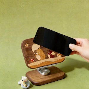 May include: A wooden phone stand featuring an inlaid design of three cartoon creatures, one holding a leaf umbrella, with a small red mushroom. The stand has a rectangular base and a swivel mount. A small white figurine sits next to the base.