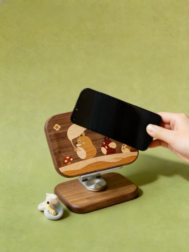 May include: A wooden phone stand featuring an inlaid design of three cartoon creatures, one holding a leaf umbrella, with a small red mushroom. The stand has a rectangular base and a swivel mount. A small white figurine sits next to the base.