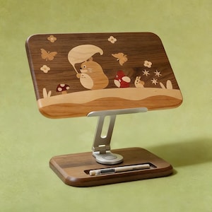 Puede incluir: Atril de madera con un diseño incrustado caprichoso que presenta un personaje de dibujos animados sosteniendo un paraguas de hojas, mariposas y flores. El soporte tiene un brazo ajustable plateado y un portalápices. La base y la parte superior son de madera clara y oscura.