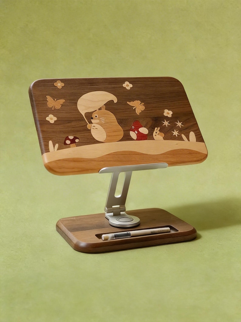 May include: A wooden tablet stand featuring a dark walnut base and a rectangular support panel with light wood inlay art depicting a creature holding a leaf umbrella, mushrooms, and butterflies. A silver metal adjustable arm connects the two parts.