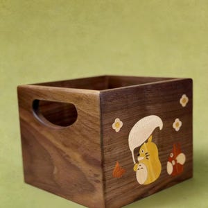 May include: A square wooden storage box with a handle cutout on one side. The box is decorated with a cartoon character design in yellow, orange, and white. The wood has a rich, dark brown color.