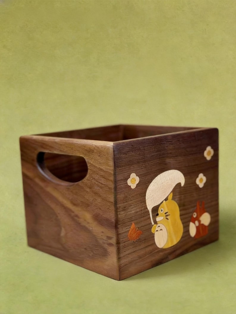 May include: A square wooden storage box with a handle cutout on one side. The box is decorated with a cartoon character design in yellow, orange, and white. The wood has a rich, dark brown color.