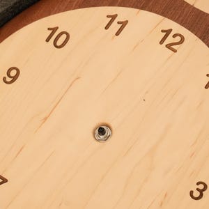 May include: A circular wall clock face crafted from light-coloured wood with a dark wood border. The face features engraved dark brown numerals from 1 to 12 arranged in a circle, with a central metal mechanism post protruding from the centre.