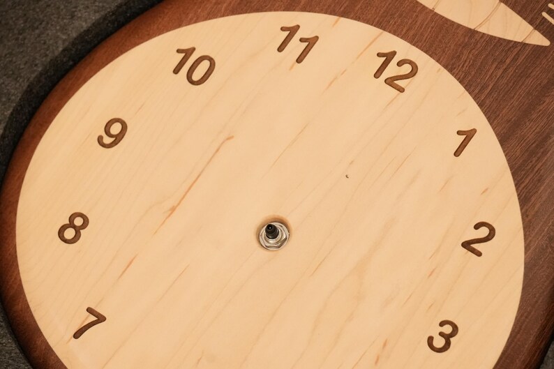 May include: A circular wall clock face crafted from light-coloured wood with a dark wood border. The face features engraved dark brown numerals from 1 to 12 arranged in a circle, with a central metal mechanism post protruding from the centre.