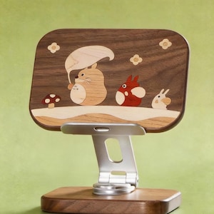 May include: A wooden phone stand featuring a dark walnut rectangular back panel with light wood inlay art depicting three cartoon creatures, a mushroom, and flowers. The stand has a silver metal adjustable arm and a matching dark walnut base.