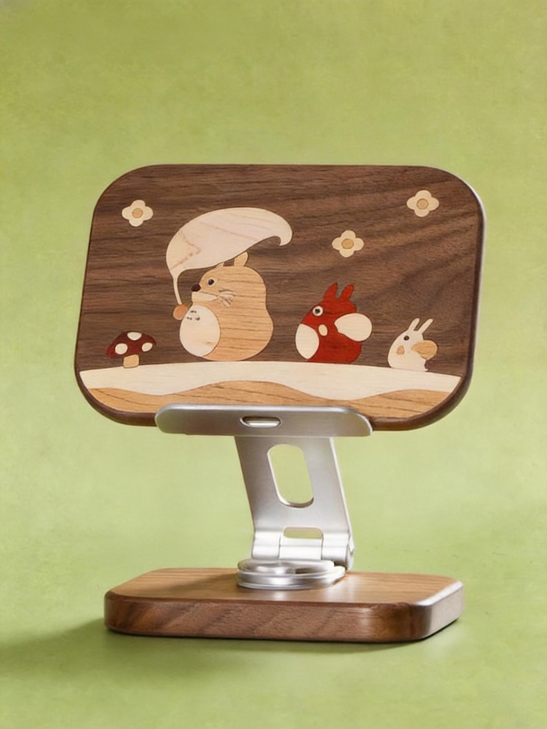 May include: A wooden phone stand featuring a dark walnut rectangular back panel with light wood inlay art depicting three cartoon creatures, a mushroom, and flowers. The stand has a silver metal adjustable arm and a matching dark walnut base.