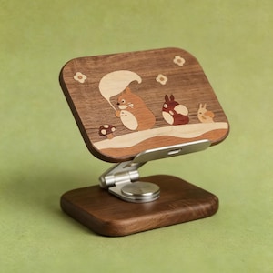 May include: A wooden phone stand featuring a rectangular dark wood panel with an inlaid design of three cartoon creatures and a mushroom. The stand has a silver metal adjustable hinge and a rectangular dark wood base for stability and support.