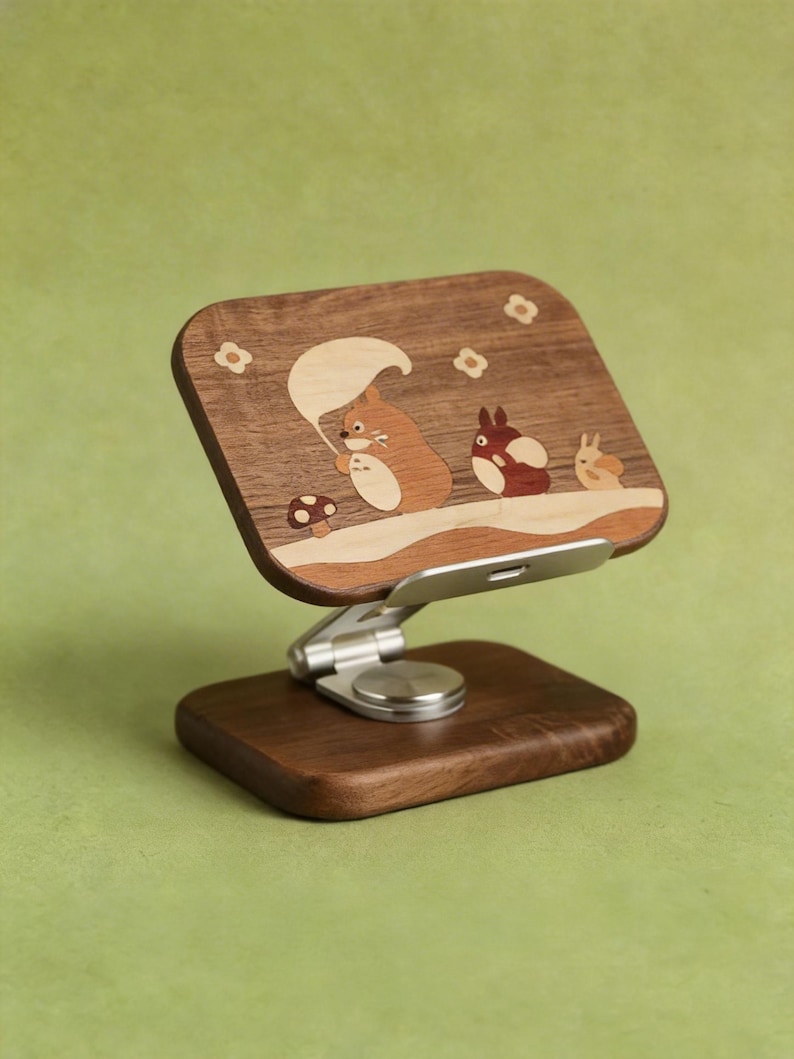 May include: A wooden phone stand featuring a rectangular dark wood panel with an inlaid design of three cartoon creatures and a mushroom. The stand has a silver metal adjustable hinge and a rectangular dark wood base for stability and support.
