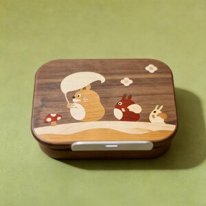 May include: A rectangular wooden bento box with rounded corners featuring an inlaid wood design on the lid. The inlay depicts three cartoon creatures, a red mushroom, and two small flowers. A light-colored horizontal latch is centered on the front.
