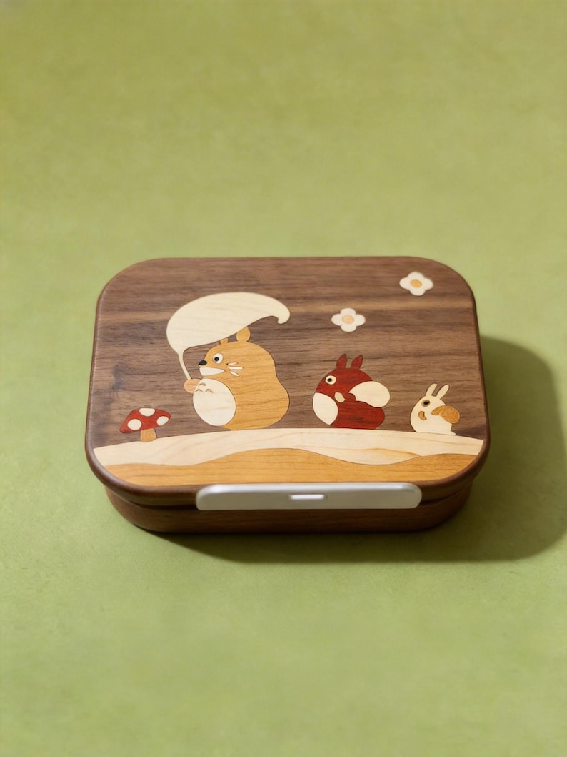 May include: A rectangular wooden bento box with rounded corners featuring an inlaid wood design on the lid. The inlay depicts three cartoon creatures, a red mushroom, and two small flowers. A light-colored horizontal latch is centered on the front.