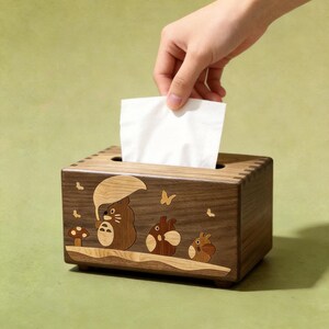 May include: A rectangular wooden tissue box holder featuring a dark brown wood grain finish. The front panel displays an inlaid wood design of three small creatures, one holding an umbrella, alongside a mushroom and butterflies.