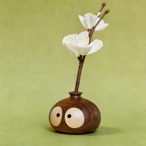 May include: A quirky wooden vase, dark brown with two large, light-coloured eye-shaped inlays. It holds a delicate stem with two white, paper-like flowers. The background is a solid, muted green.