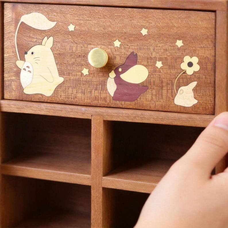 K&ouml;nnte beinhalten: Holzschrank mit einer Schublade mit eingelegten Cartoon-Figuren und Sternen. Die Schublade hat einen Messingknopf und ist mit einer hellbraunen und dunkelbraunen kaninchenartigen Figur mit einem Blatt verziert. Der Schrank hat mehrere offene F&auml;cher.